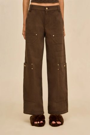 Lower torso and legs of a person wearing brown cargo pants with large side pockets and metal rivets, standing in brown fuzzy slippers.