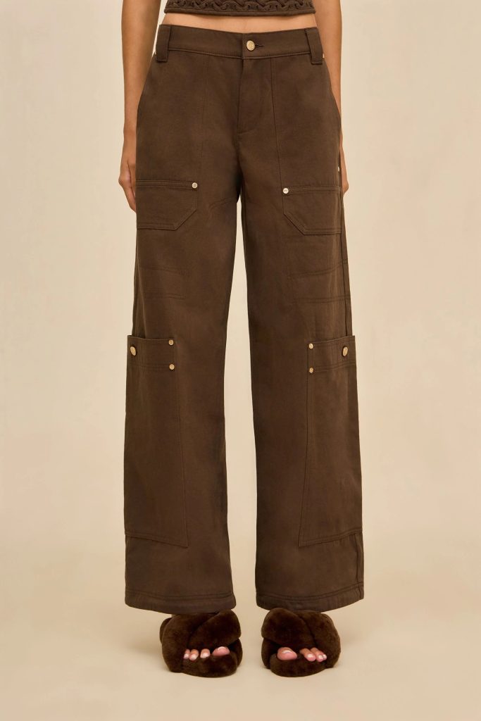 Lower torso and legs of a person wearing brown cargo pants with large side pockets and metal rivets, standing in brown fuzzy slippers.