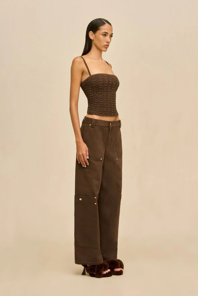 Model posing in a brown crochet crop top with spaghetti straps and brown cargo pants, wearing fuzzy brown slippers against a beige backdrop.