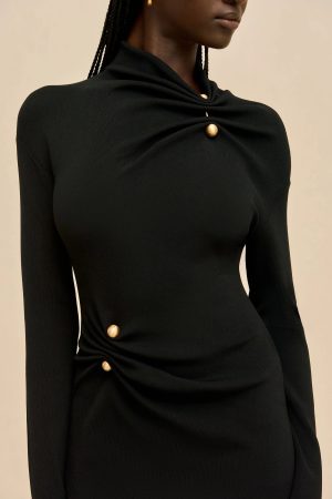 Close-up of a person wearing a black knit top with a twisted knot and gold button accents at the chest against a beige background.