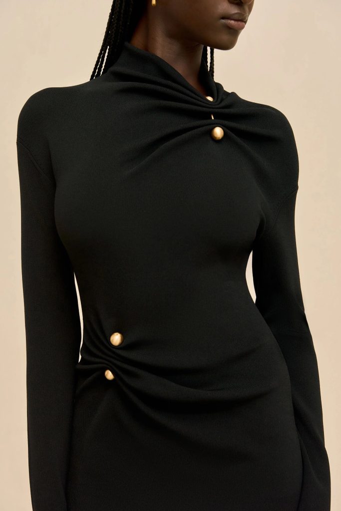 Close-up of a person wearing a black knit top with a twisted knot and gold button accents at the chest against a beige background.