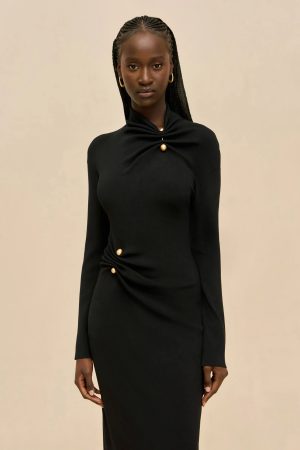 Portrait of a young Black woman with braided hair wearing a black long-sleeve dress with gold buttons, standing against a beige wall.