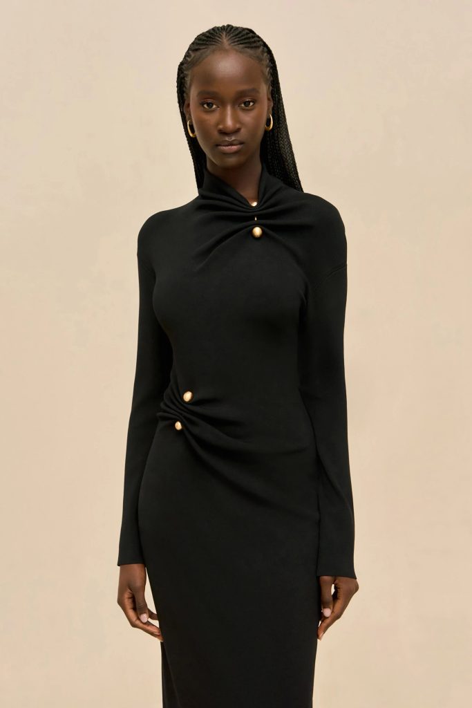 Portrait of a young Black woman with braided hair wearing a black long-sleeve dress with gold buttons, standing against a beige wall.