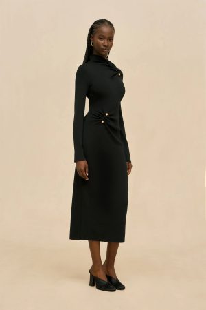 Full-body portrait of a woman wearing a long black dress with gold buttons, standing against a beige backdrop, wearing black heeled mules and gold hoop earrings.
