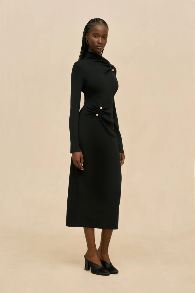 Full-body portrait of a woman wearing a long black dress with gold buttons, standing against a beige backdrop, wearing black heeled mules and gold hoop earrings.