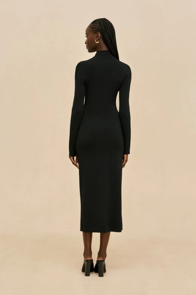 Back view of a woman in a long black dress with braided hair and a gold hoop earring, standing against a beige background.