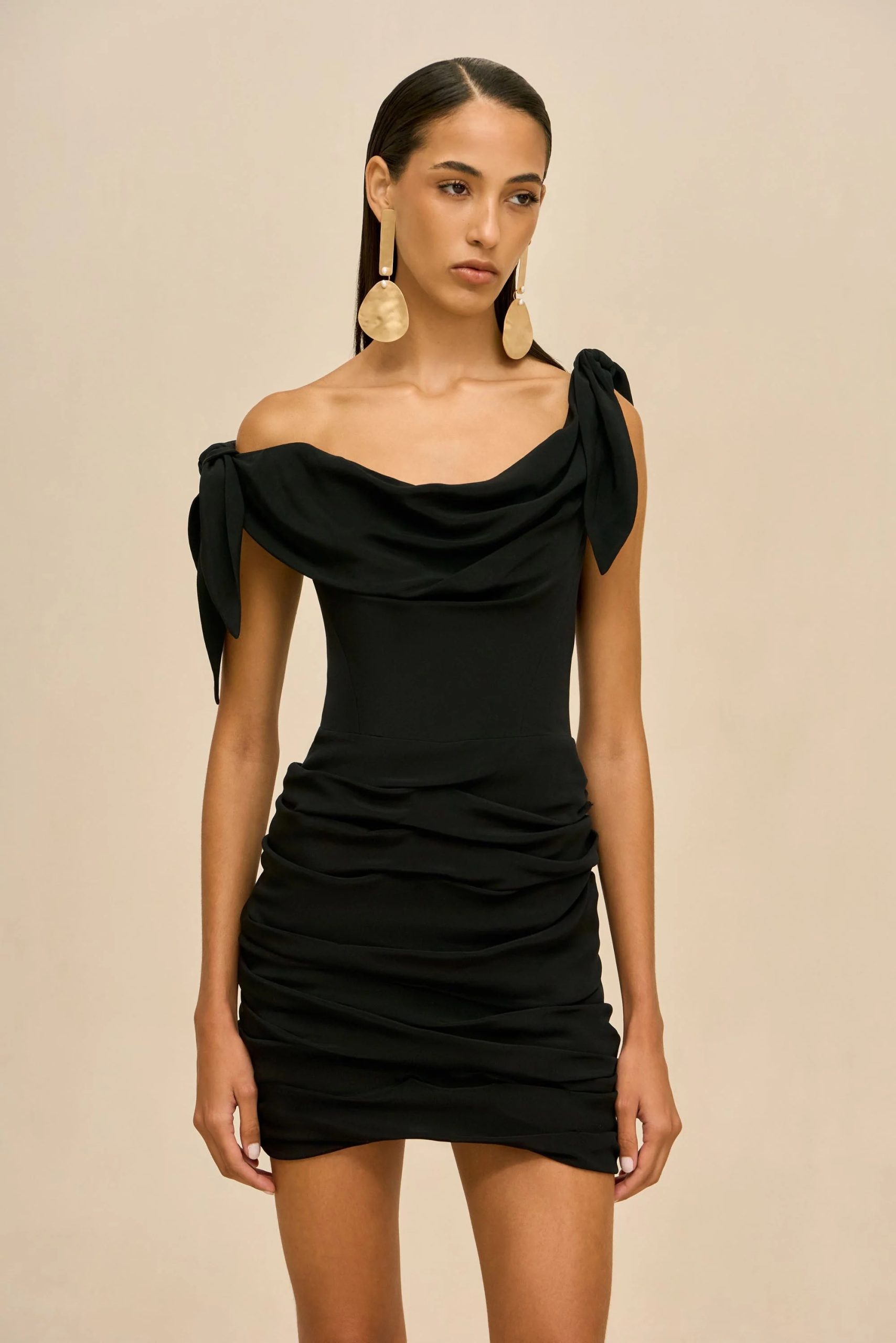 Image 506 - New Trend Model wearing a black off-shoulder draped dress with large gold teardrop earrings against a beige backdrop.