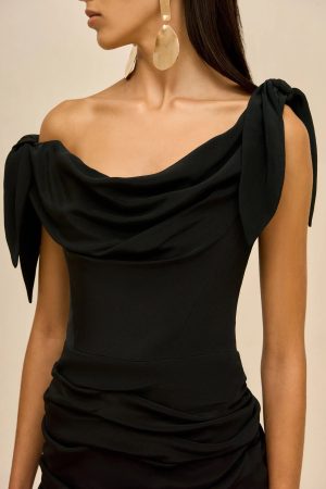 Close-up of a woman wearing a black draped off-shoulder dress and large gold teardrop earrings against a beige background, shown in profile.