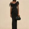 Woman in a black off-the-shoulder evening gown with draped fabric, posing against a beige backdrop, holding a black fringe clutch.