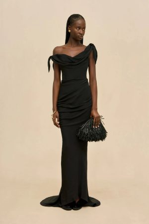 Woman in a black off-the-shoulder evening gown with draped fabric, posing against a beige backdrop, holding a black fringe clutch.