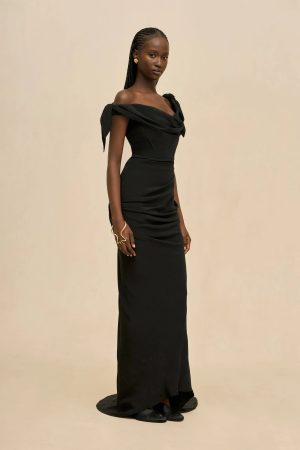 Woman in a fitted black off-shoulder evening gown with a draped neckline, standing against a beige backdrop, wearing gold earrings and braided hair.