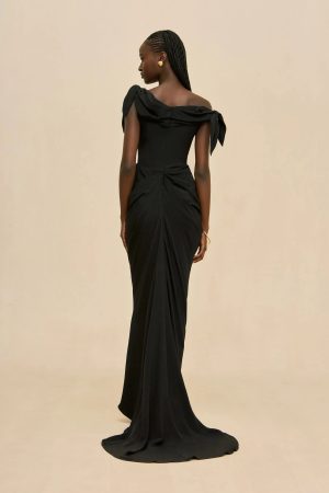 Back view of a woman in a black off-the-shoulder evening gown with draped shoulders and a long train, against a beige backdrop.