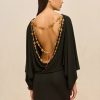 Side profile of a woman in a black backless dress wearing layered beaded necklaces and a gold hoop earring against a beige backdrop.