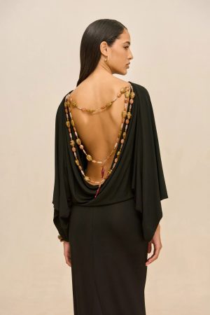 Side profile of a woman in a black backless dress wearing layered beaded necklaces and a gold hoop earring against a beige backdrop.