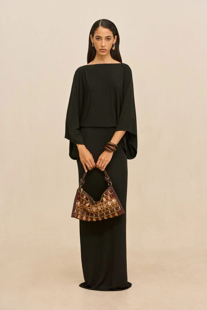 Woman in a long black dress with voluminous sleeves stands against a beige backdrop, holding a patterned beaded handbag at waist level.