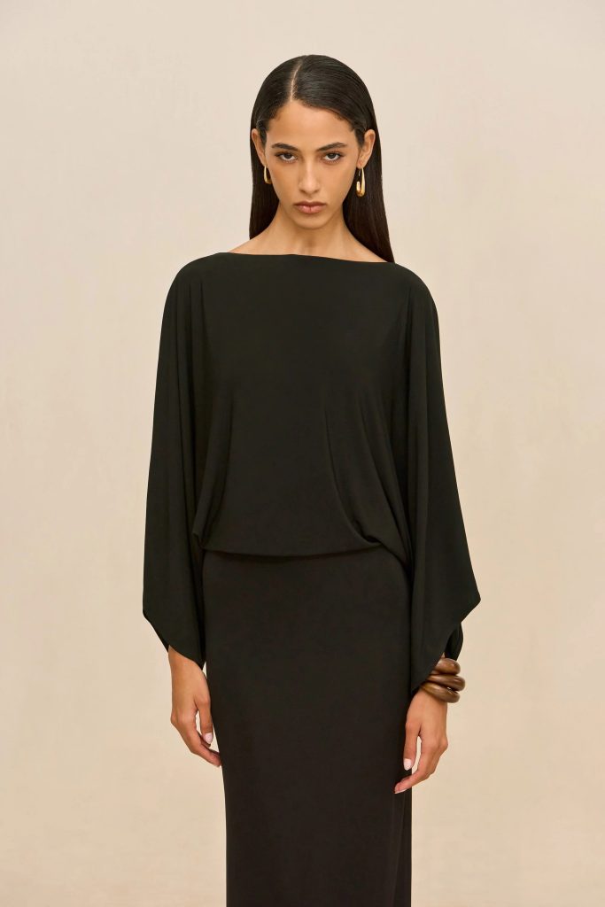 Female model in a black long-sleeve dress with draped sleeves and gold hoop earrings against a beige backdrop.
