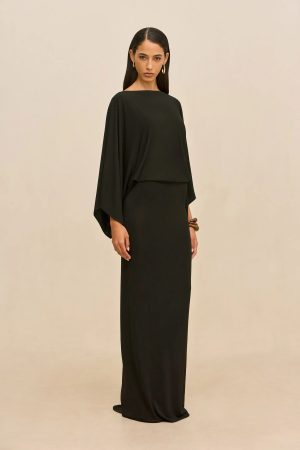 Model wearing a black, one-shoulder draped gown, standing against a beige backdrop with gold hoop earrings.
