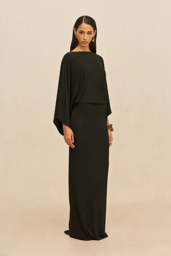 Model wearing a black, one-shoulder draped gown, standing against a beige backdrop with gold hoop earrings.