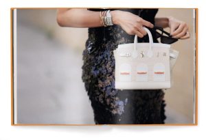 Woman in a dark sequined dress holds a white handbag with orange accents, displayed in an open fashion book