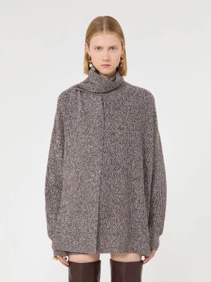 Woman wearing an oversized gray knit turtleneck sweater, standing against a white backdrop, looking at the camera.