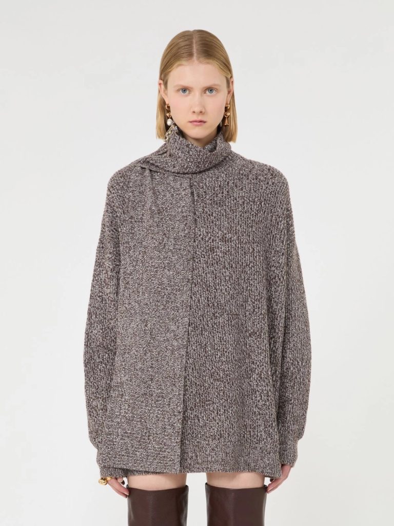 Woman wearing an oversized gray knit turtleneck sweater, standing against a white backdrop, looking at the camera.
