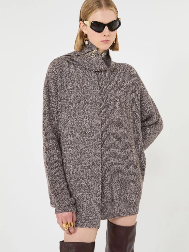 Model wearing a chunky, speckled brown knit turtleneck sweater dress with black sunglasses and gold jewelry, paired with tall boots.