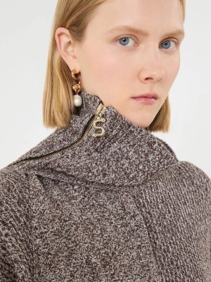 Close-up of a woman with blond hair wearing a brown knit turtleneck and gold jewelry, including a hoop earring with a dangling pearl.