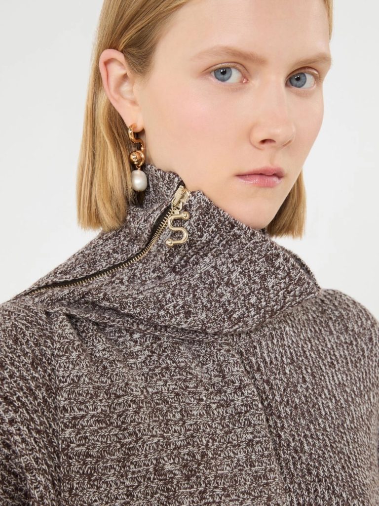 Close-up of a woman with blond hair wearing a brown knit turtleneck and gold jewelry, including a hoop earring with a dangling pearl.