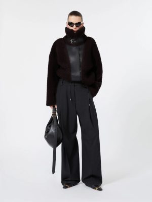 Model in oversized black shearling jacket with high collar, black wide-leg trousers, and black sunglasses, holding a black bag.
