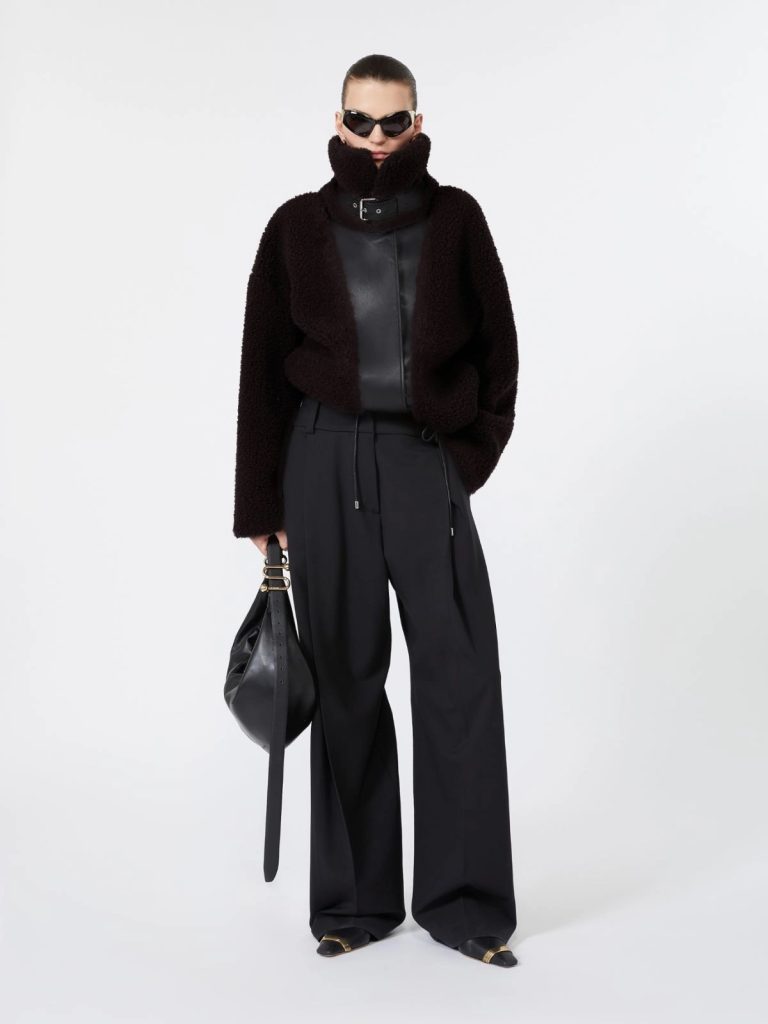 Model in oversized black shearling jacket with high collar, black wide-leg trousers, and black sunglasses, holding a black bag.
