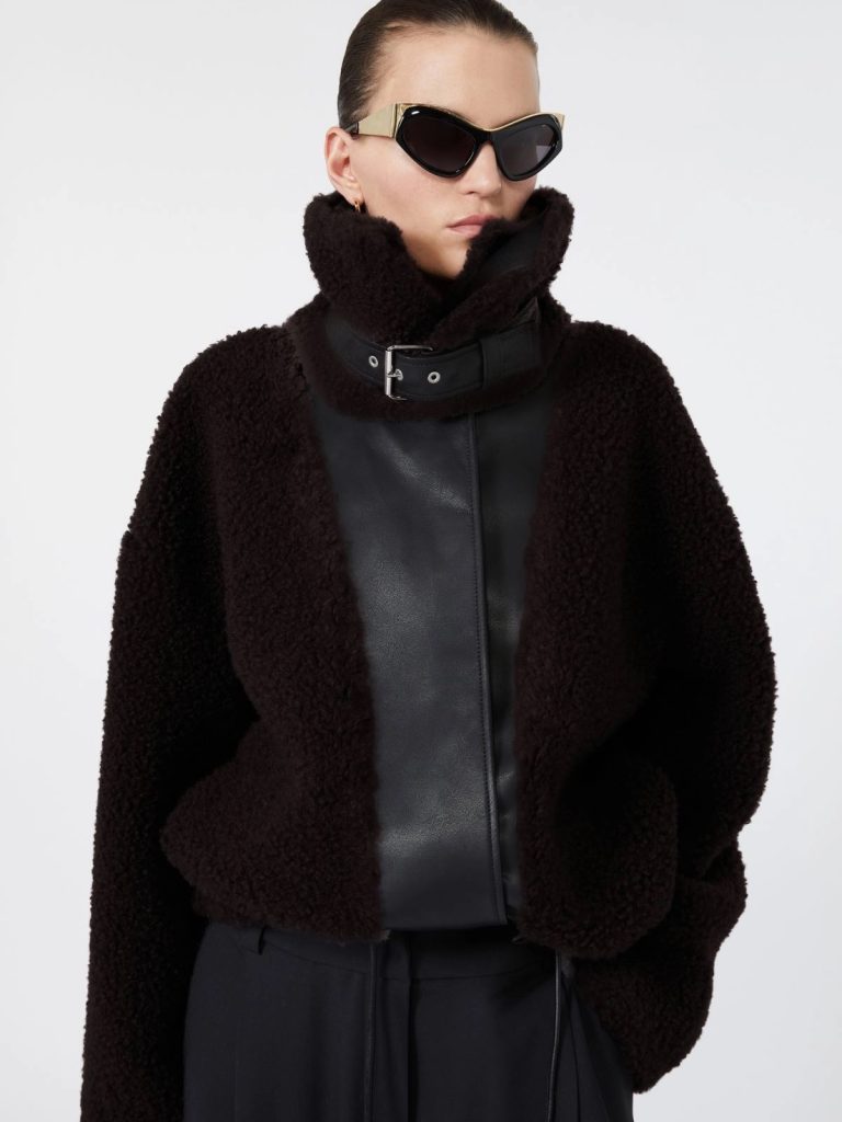 Model wearing an oversized black shearling coat with a leather front panel and a high collar, plus black sunglasses with gold accents.