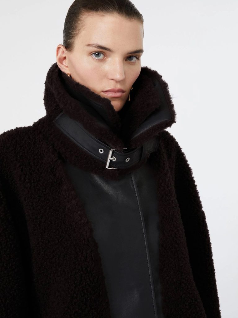 Close-up of a woman wearing a black shearling coat with a high collar and buckle detail, neutral expression.