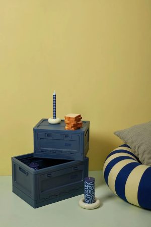 Blue stacking storage crates with a tall blue-and-white dotted candle upright on a white base and a small stack of toast on top, set against a pale yellow wall with a striped blue and white cushion nearby.