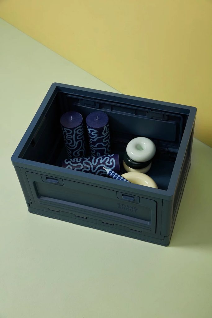 Open dark blue storage box containing two tall patterned blue candles and white ceramic bowls on a pale yellow-green surface.
