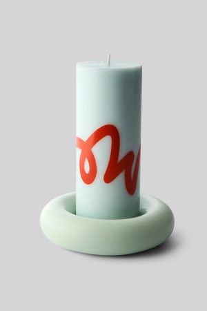 Tall pale blue pillar candle with a red decorative script on its side, placed on a rounded pale ring stand against a gray background.