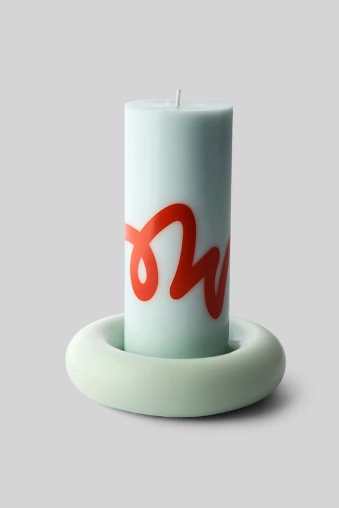 Tall pale blue pillar candle with a red decorative script on its side, placed on a rounded pale ring stand against a gray background.