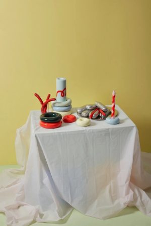 Still life on a white-draped table: colorful glossy ceramic shapes (rings, donuts) and candles against a yellow background.