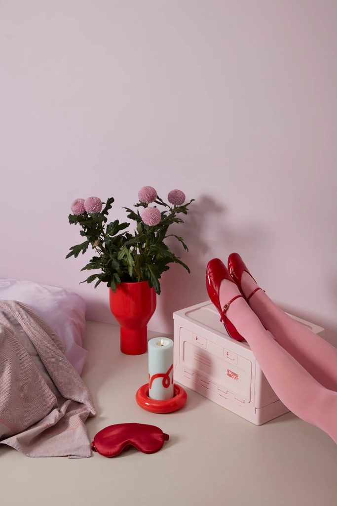 Pink-themed still life: red vase with pink flowers, red shoes on a white box, candle, eye mask, and soft fabric on a pale surface.