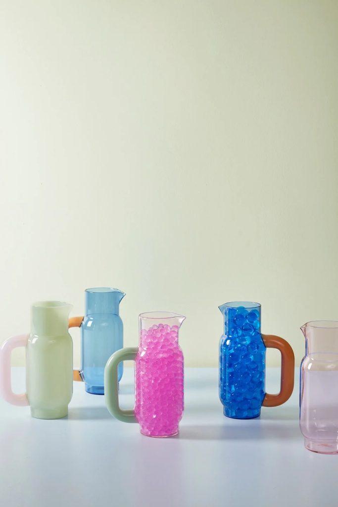 Row of pastel glass pitchers on a white table, some filled with pink or blue beads.