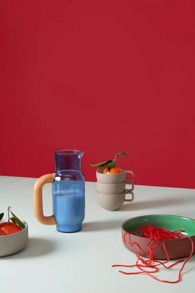 Blue pitcher with orange handle on a white table, beside a stack of beige mugs with an orange and leaves, and a bowl with red string against a red wall.