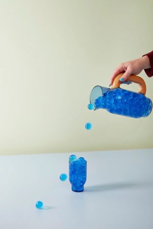 Hand pours bright blue beads from a transparent pitcher with an orange handle into a blue cup on a pale table, with several beads midair and on the surface.