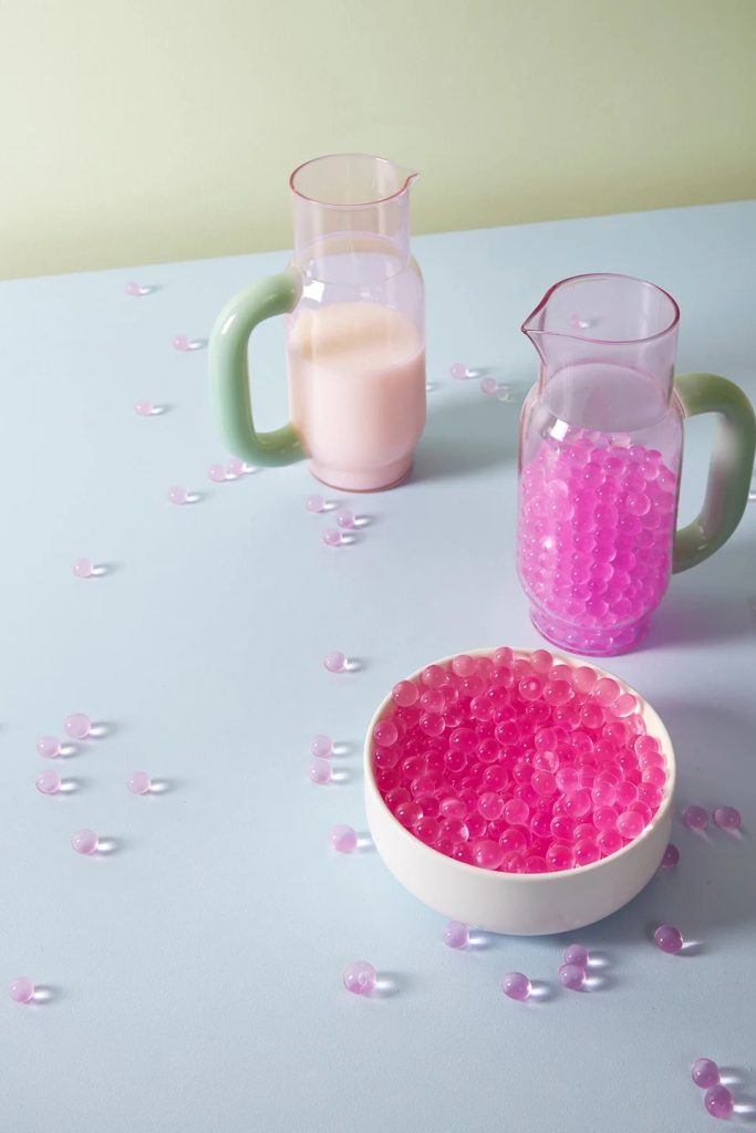 Pastel still life: two glass pitchers (one with pink liquid, one with beads) and a white bowl of pink beads on a light blue surface, with scattered beads around.