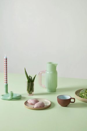 Pastel still-life on a light green table: mint-green pitcher, pink candle, small plant in a glass, brown cup, plate with two pink pastries, and lettuce.