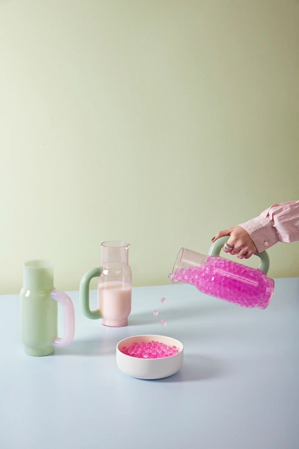 Image 584 - New Trend Hand holds a pink-bead pitcher pouring beads into a white bowl on a light blue table, pastel backdrop in view.