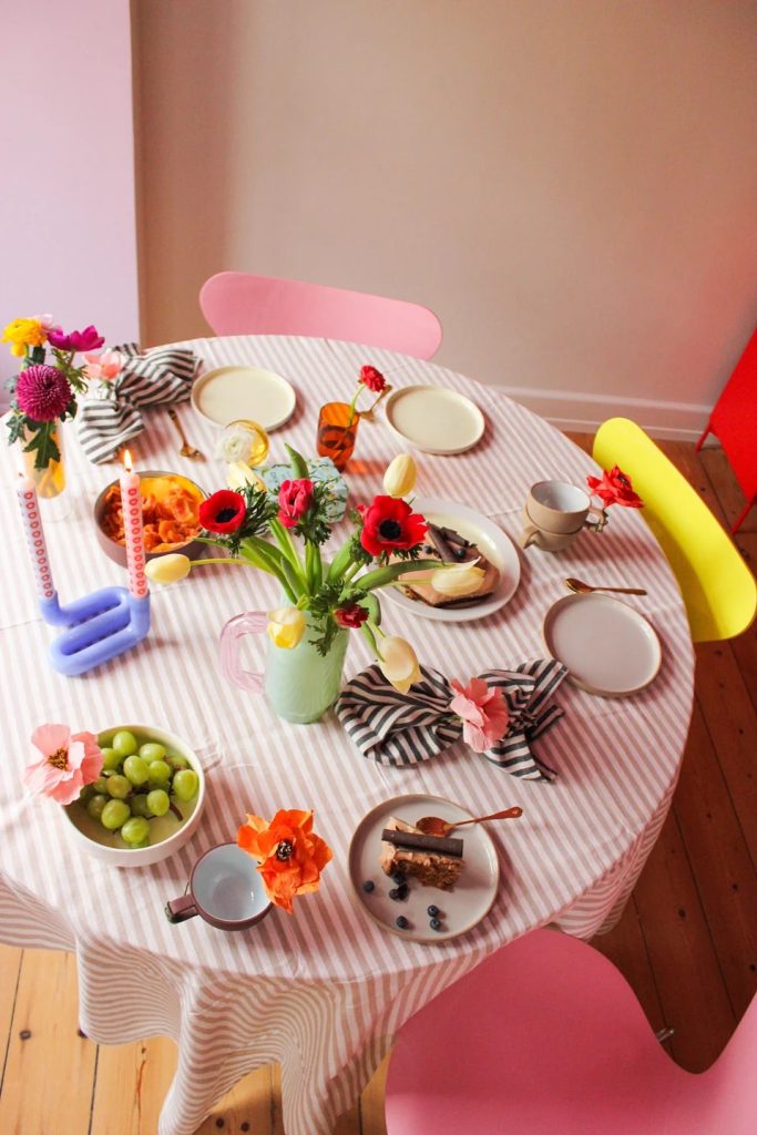 Colorful breakfast table set with flowers, candles, and pastel chairs; plates with food and a striped tablecloth, pink and yellow accents.