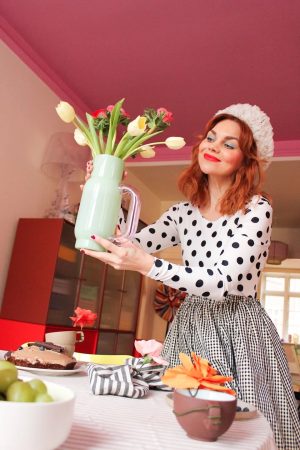 Woman with red hair in a polka-dot top and gingham skirt holds a pastel green vase of tulips over a decorated party table.