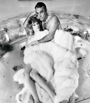 Couple lying in a large glass bathtub, wrapped in a white fur rug, black-and-white vintage photo.
