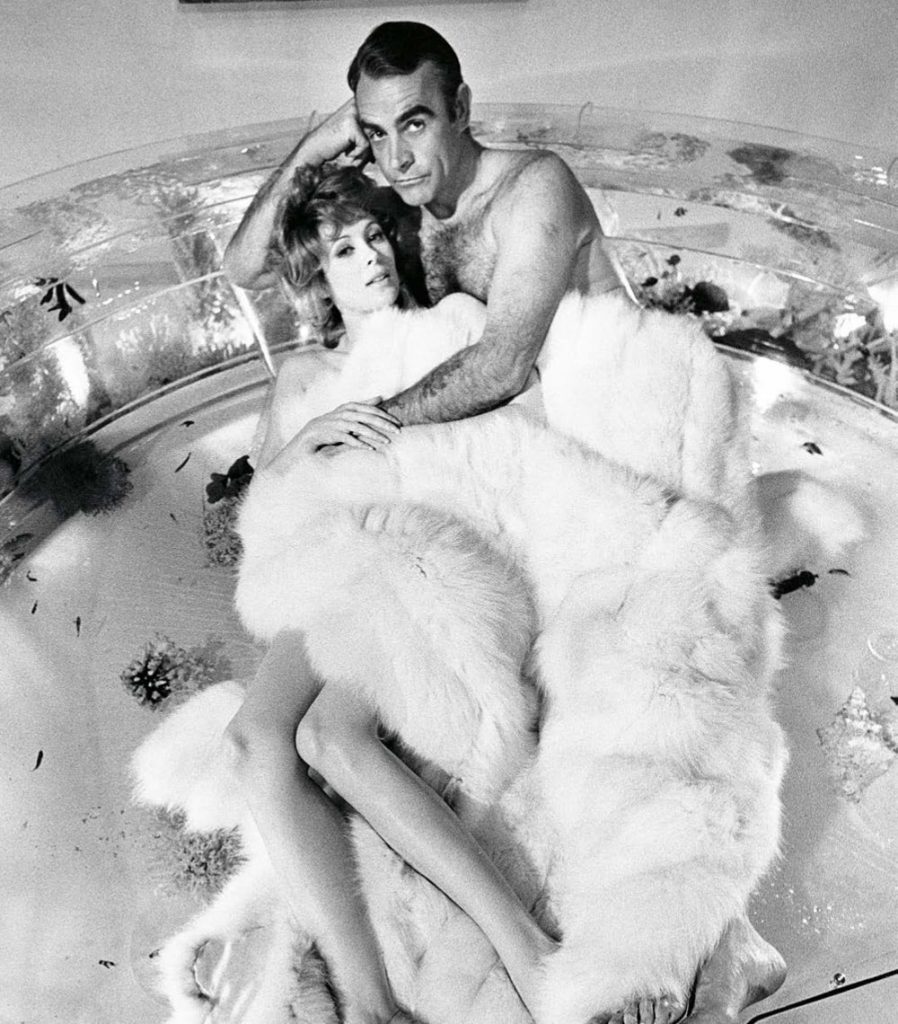 Couple lying in a large glass bathtub, wrapped in a white fur rug, black-and-white vintage photo.