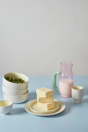 Still life with three white bowls filled with peas, a plate with butter blocks, a pink pitcher of milk, and two white cups on a pale blue surface.