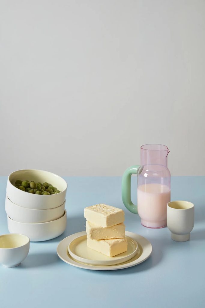 Still life with three white bowls filled with peas, a plate with butter blocks, a pink pitcher of milk, and two white cups on a pale blue surface.